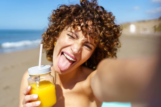 Playful female with ginger curly hair showing tongue while holding glass cup of orange juice and taking selfie on coast