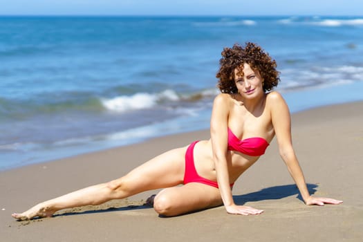 Full body of slim female with red curly hair in bikini looking at camera while lying on sandy coast of blue waving sea