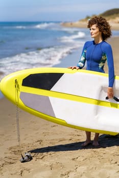 Side view of smiling young female redhead in swimsuit standing on sandy beach with paddleboard in hands and waiting while looking away against blurred seacoast