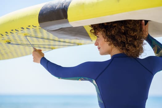 Back view of positive curly haired female in trendy swimwear with colorful paddleboard on background of sea looking away in bright daylight