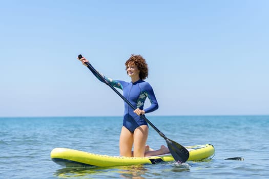 Full body view of young female with curly hair in wetsuit kneeling on paddleboard on blue sea while paddling and looking away