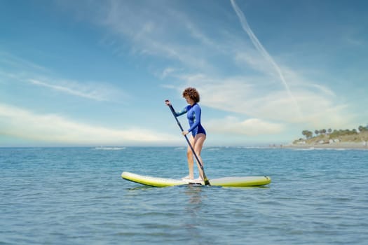 Focused female surfer in blue wetsuit standing on SUP board and paddling in sea against cloudy blue sky in sunny summer day