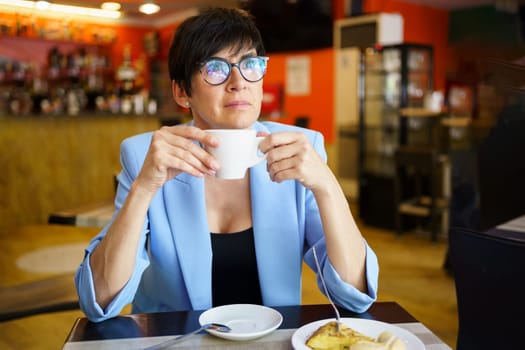 Thoughtful middle aged female in blue jacket and eyeglasses sitting at table with cup of hot beverage and looking away while resting in cafe