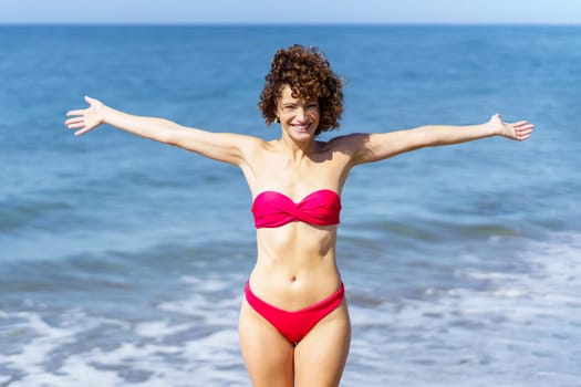 Cheerful young curly ginger haired female in pink bikini with outstretched hands and looking at camera, while standing near foamy rippling seawater against waving sea and blue sky