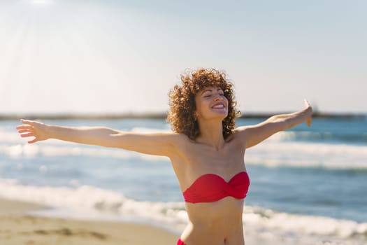 Cheerful young female redhead in pink bikini with eyes closed chin up and smiling with outstretched arms, in sunlight while standing on sandy beach against blue seawater and sky