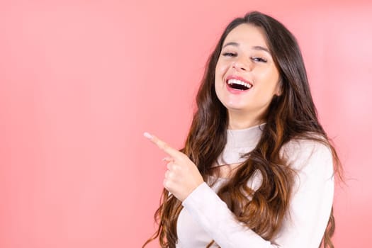 Long-haired woman with shocked and amused expression points finger to new visitor in pink studio. Lady smiles covering mouth at pink studio wall
