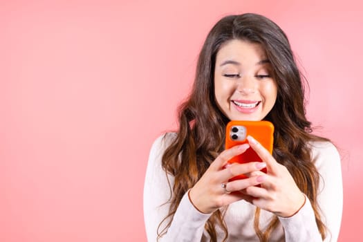 Brown-haired young woman scrolls social media humming favourite music at pink wall studio. Lady scrolling news reads with delighted expression