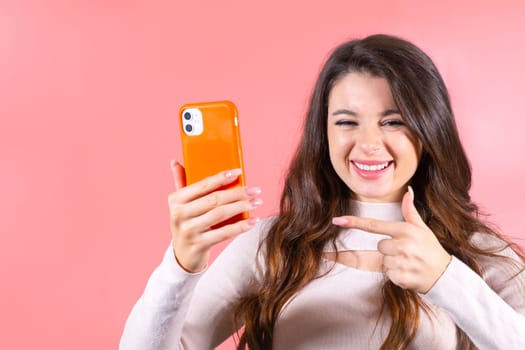 Young woman with delighted expression with natural makeup pointing at her smartphone.