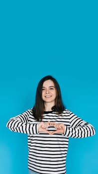 Joyful Woman in Striped Sweater Smiling, Gesturing Heart Hands - Symbol of Love and Happiness - Isolated on Blue Background - Perfect for Positive Emotions, Love, and Lifestyle Concepts.