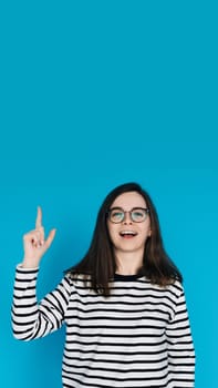 Excited Cheerful Woman in Striped Sweater with Glasses Pointing Up - Energetic Female Model Gesturing towards Empty Space - Isolated on Blue Background - Perfect for Advertising, Promotions Concepts