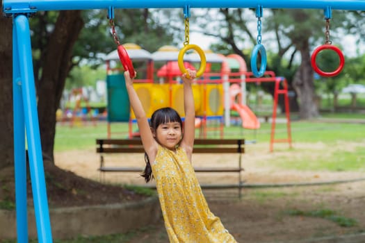 Happy girl hanging on monkey bar by hand doing exercise. Little Asian girl playing at outdoor playground in the park on summer vacation. Healthy activity.