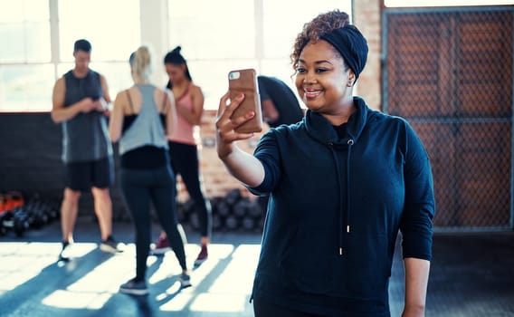 Soon Ill be changed. a cheerful young woman standing and taking a self portrait with her cellphone before a workout in a gym