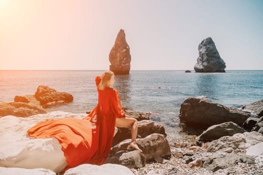 Woman travel sea. Young Happy woman in a long red dress posing on a beach near the sea on background of volcanic rocks, like in Iceland, sharing travel adventure journey