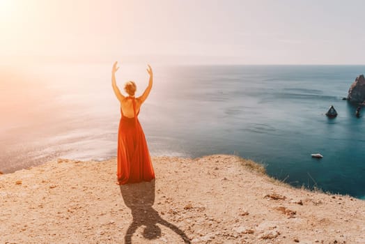 Side view a Young beautiful sensual woman in a red long dress posing on a rock high above the sea during sunrise. Girl on the nature on blue sky background. Fashion photo.