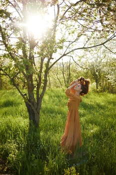 a beautiful, joyful woman stands in a long orange dress, in the countryside, near a tree blooming with white flowers, during sunset, illuminated from behind and touches her long hair. High quality photo