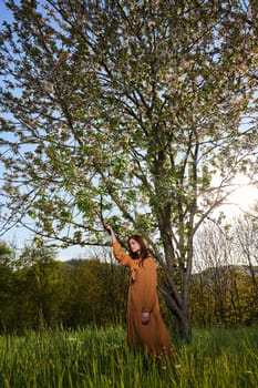 a sweet, attractive woman with long red hair is standing in the countryside near a flowering tree in a long orange dress and touching a branch with her hand looks at the camera. High quality photo