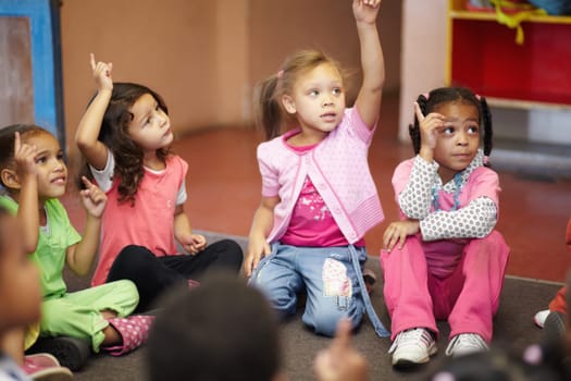 Education, kindergarten and kids asking a question with hands raised while sitting on a classroom floor for child development. School, learning or curiosity with children in class to study for growth.