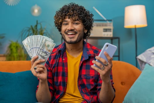 Planning family budget. Smiling indian man counting money cash, use smartphone calculate domestic bills at home room. Joyful hindu guy satisfied of income and saves money for planned vacation, gifts