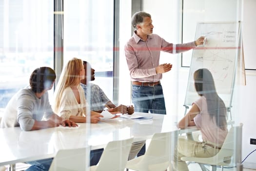 Planning, mature mentor with colleagues and chart in a boardroom of their modern workplace. Teamwork or collaboration, data review or brainstorming and coworkers working together in business meeting.