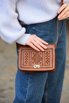 a red-haired girl in a blue jeans and a sweater poses outside with a small leather handbag.