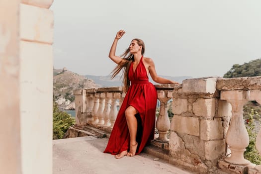 Side view a Young beautiful sensual woman in a red long dress posing on a volcanic rock high above the sea during sunset. Girl on the nature on overcast sky background. Fashion photo