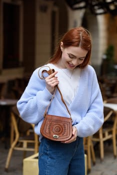 a red-haired girl in a blue jeans and a sweater poses outside with a small leather handbag.