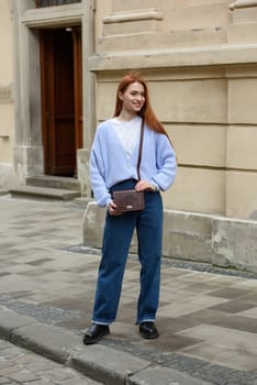 a red-haired girl in a blue jeans and a sweater poses outside with a small leather handbag.