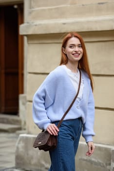 a red-haired girl in a blue jeans and a sweater poses outside with a small leather handbag.