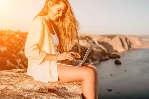 Successful business woman in yellow hat working on laptop by the sea. Pretty lady typing on computer at summer day outdoors. Freelance, travel and holidays concept.