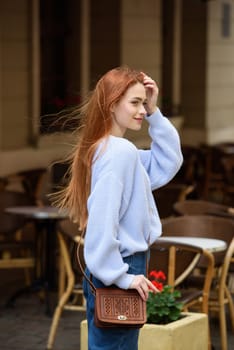 a red-haired girl in a blue jeans and a sweater poses outside with a small leather handbag.