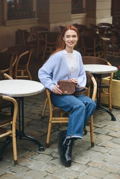 a red-haired girl in a blue jeans and a sweater poses outside with a small leather handbag.