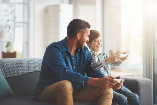 Youre getting good. a cheerful little boy and his father playing video games together on the television while being seated on the sofa at home during the day