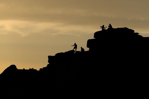 People on stanage edge, peak district, at sunset. A couple take a selfie holding drinks, silhouetted against the golden sky. Climbers stand there too