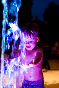 The child is frolicking in the fountain in the park, he is very happy. Have fun and enjoy the summer evening. Walking and recreation. Portrait of a happy child. Night shooting