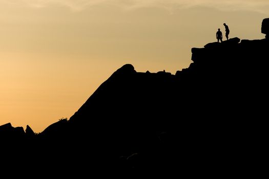 A pair of rock climbers, silhouetted at sunset, look back from the summit of stange edge. Located in the peak district in england. Outdoor activity