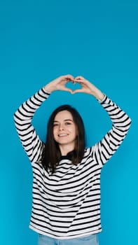Joyful Woman in Striped Sweater Expressing Love and Happiness - Happy Girl Showing Heart Gesture with Arms, Smiling - Isolated Blue Background with Copy Space - Perfect for Love, Happiness Concepts