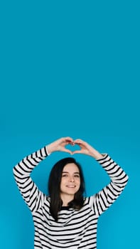 Joyful Woman in Striped Sweater Spreading Love - Smiling Girl with Raised Arms Creating Heart Shape, Radiating Happiness and Positivity - Isolated Blue Background. Perfect for Positive Vibes Concepts