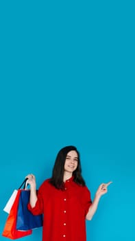 Cute Woman in Red Shirt with Shopping Bag - Pointing to Empty Space - Perfect for Promoting Sales and Discounts - Isolated on Blue Background. Smiling Woman with Shopping Bag