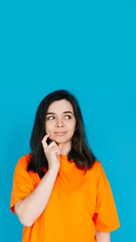 Attractive Young Woman in Trendy Orange Clothes, Pensive and Skeptical Expression, Empty Space for Copy, Isolated on Blue Background - Portrait Photo.