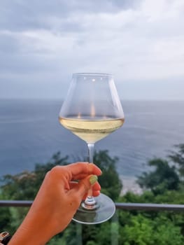 During sunset a female hand toasts the new year with a glass of white wine. Photo taken on a balcony of a cruise ship with a sea view.