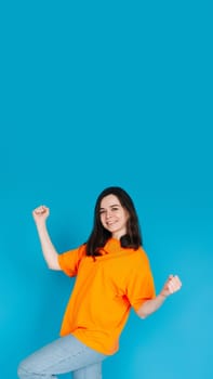 Excited Young Woman Celebrating Huge Sales Promotion, Raising Fists in Victory, Trendy Orange Outfit, Isolated on Blue Background, Portrait Photo.