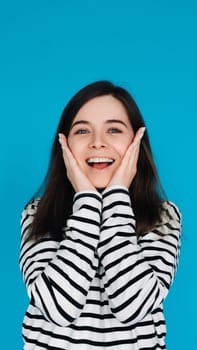 Expressive Portrait of a Surprised and Joyful Young Woman with Palms on Cheekbones, Isolated on Blue Background.