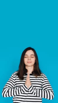 Serene Portrait of a Beautiful Young Woman Meditating with Closed Eyes and Palms, Inner Peace and Relaxation, Isolated on Blue Background.