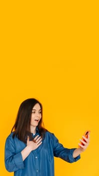 Digital Surprise: Captivating Young Woman in Stylish Shirt, Reading Astonishing News on her Phone - Vibrant Expression Isolated on Yellow Background