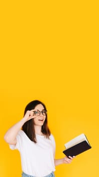 Portrait of a happy young woman wearing glasses and white T-shirt, immersed in reading a book with an open mouth, isolated on a yellow background.