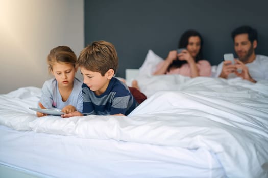 They still love chilling on Mom and Dads bed. an adorable little girl and boy using a tablet together while their parents are chilling in bed at home