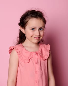 Vertical studio portrait of Caucasian blue eyed smiling coquettish little girl 5-6 years old, wearing summer pink dress, looking at camera, isolated on pink color background. Kids fashion and beauty