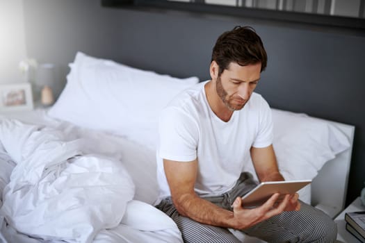 I always check my messages before I get up. a handsome middle aged man using his digital tablet while relaxing in his bedroom