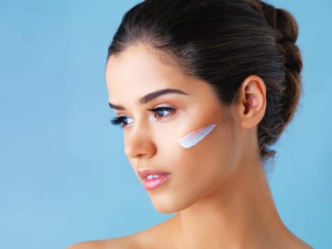 The secret to her perfect skin. Studio shot of a beautiful young woman with lotion on her face against a blue background