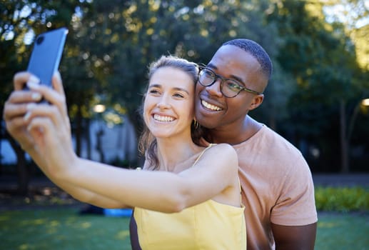 Selfie, phone and interracial couple smile in park enjoying weekend, summer holiday and quality time together. Nature, dating and happy man and woman relax, calm and hug to take picture on smartphone.
